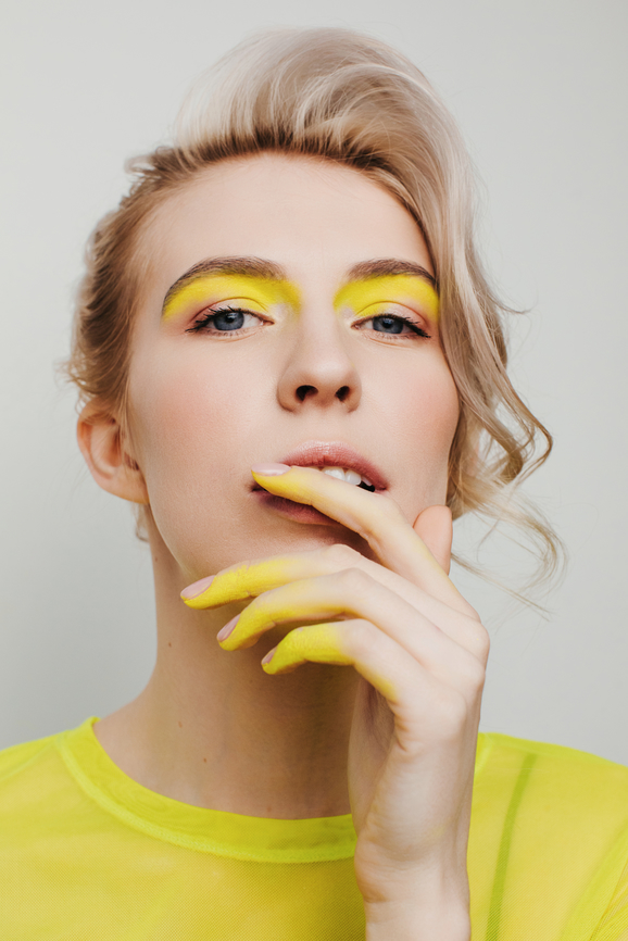 Eine blonde Frau mit einem gelben Shirt fasst sich mit ihren gelb geschminkten Fingern an den Mund.