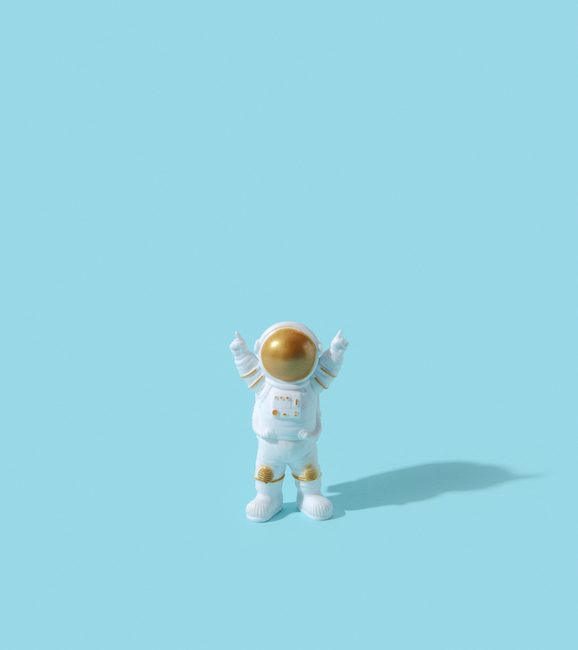 Happy toy astronaut standing over blue background with raised hands. Cosmonaut celebrating success during expedition. Human achievements.